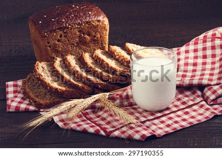 Fresh baked loaf and slices of wholegrain rye bread with spikelets of wheat and glass of milk on a red and white cloth on a dark wooden boards. close up