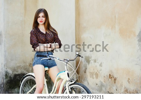 Attractive young woman with long fair hair wearing on blouse and shorts is posing on the bicycle on the old wall background, waist up