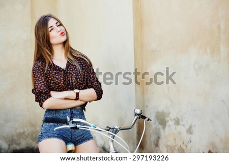 Cheerful girl with long fair hair wearing on blouse and shorts is posing on the bicycle on the old wall background, waist up