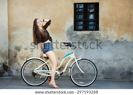 Young girl with long fair hair wearing on dark blouse and blue shorts is posing on the bicycle on the old wall background, on the street of European city