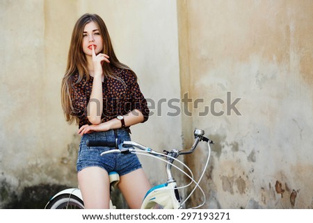Passionate young woman with long straight fair hair wearing on dark blouse and blue shorts is posing on the bicycle on the old wall background
