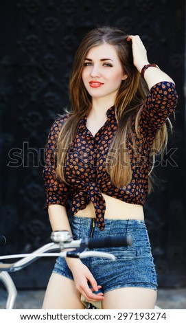 Charming young woman with long straight fair hair wearing on dark blouse and blue shorts is sitting on the bicycle and looking at camera on the street of old European city