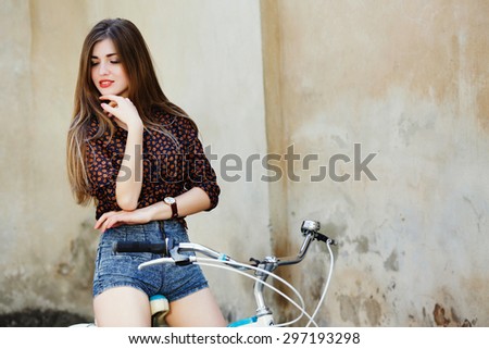 Adorable girl with long straight fair hair wearing on dark blouse and blue shorts is posing on the bicycle on the old wall background
