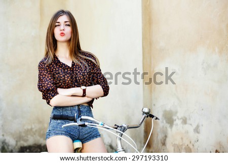 Charming girl with long fair hair wearing on short blouse and shorts is posing on the bicycle on the old wall background, waist up