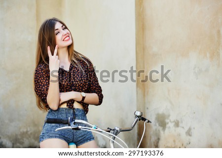 Cheerful girl with long fair hair wearing on dark blouse and shorts is posing on the bicycle on the old wall background, in  the European city, waist up