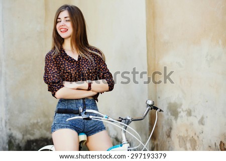 Positive young woman with long fair hair wearing on dark blouse and shorts is posing on the bicycle on the old wall background, waist up