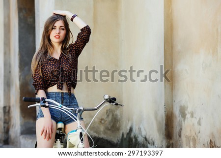 Charming young woman on dark blouse and blue shorts is sitting on the bicycle and looking at camera on the old wall background