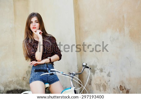 Nice girl with long straight fair hair wearing on dark blouse and blue shorts is posing on the bicycle on the old wall background in the European city