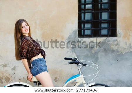Lovely girl with long fair hair wearing on dark blouse and blue shorts is sitting on the bicycle on the old wall background