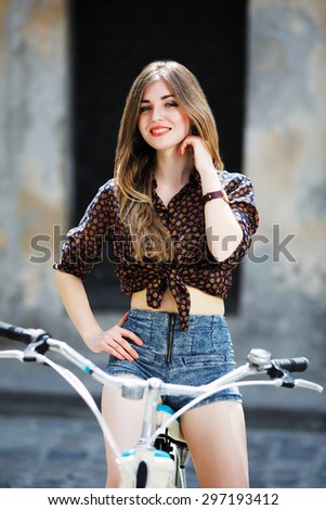 Smiling pretty girl wearing on dark blouse and blue shorts with long straight fair hair is posing on the bicycle on the street of old city