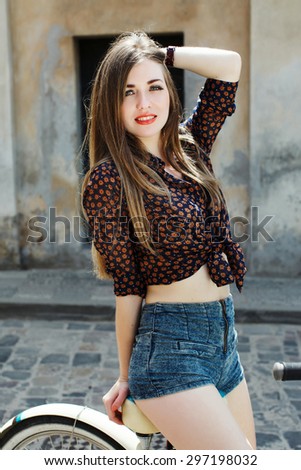 Beautiful girl wearing on dark blouse and blue shorts with long straight hair sitting on bicycle and looking at camera on the street of old European city