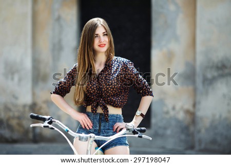 Young nice girl wearing on dark blouse and blue shorts with long straight fair hair is posing on the bicycle on the street of old European city