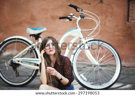 Adorable young girl with long hair wearing on dark blouse and blue shorts sitting on tiled pavement and holding her mobile phone with bicycle on a background on the street of old city