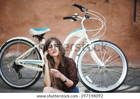 Pretty girl with long hair wearing on dark blouse and shorts sitting on tiled pavement and holding her mobile phone with bicycle on a background on the street of old city