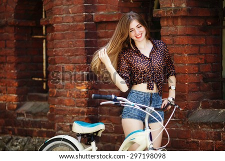 Cute smiling girl with long hair wearing on dark blouse and shorts with bicycle standing near the brick wall on the street of old city