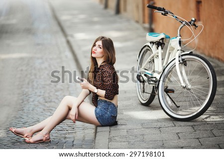 Young girl wearing on dark blouse and blue shorts with long fair hair sitting on tiled pavement near the bicycle and holding her mobile phone on the street of old city