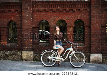 Smiling young girl with long blond hair wearing on dark blouse and blue shorts posing with bicycle near the brick building, on the old city street