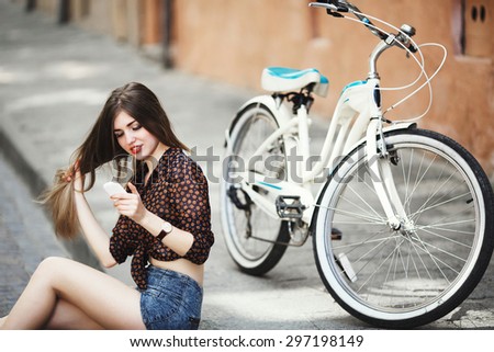 Gorgeous girl with long hair wearing on dark blouse and blue shorts sitting on tiled pavement near the bicycle and looking at her mobile phone on the street of old city