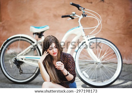 Beautiful girl with long hair wearing on dark blouse and blue shorts sitting on tiled pavement and looking at her mobile phone with bicycle on a background on the street of old city