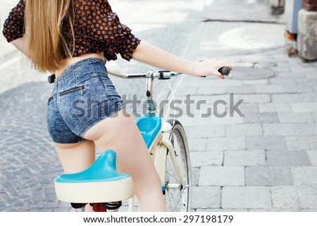Rear view of young girl which wearing on dark blouse and blue shorts with long fair hair is sitting on bicycle on the street of old European city