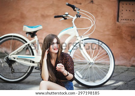 Beautiful young girl with long straight hair wearing on dark blouse and blue shorts sitting on tiled pavement and holding her mobile phone with bicycle on a background on the street of old city