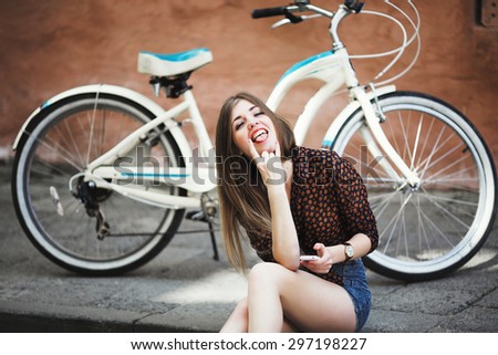 Cheerful young girl with long hair wearing on blouse and shorts sitting on tiled pavement and holding her mobile phone with bicycle on a background on the street of old city