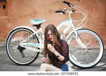 Young girl with long fair hair wearing on short dark blouse and shorts sitting on tiled pavement and looking at her mobile phone with bicycle on a background on the street of old city