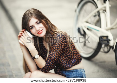 Nice young woman with long straight hair wearing on short dark blouse is posing with coffee on tiled pavement with bicycle on a background on the street of old city