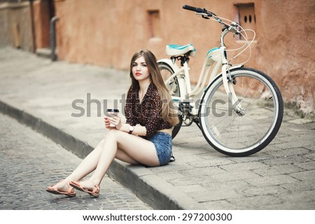 Beautiful young woman with long straight hair wearing on dark blouse and shorts is posing with coffee on tiled pavement with bicycle on a background on the street of old European city