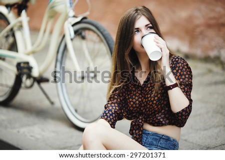 Pretty girl with long blond hair wearing on blouse and shorts is drinking coffee on tiled pavement with bicycle on a background in old city center