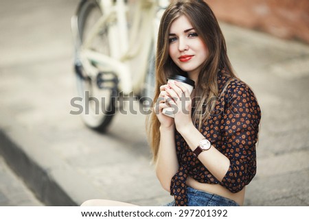 Young girl with fair hair wearing on short dark blouse is posing with coffee on tiled pavement with bicycle on a background on the street of old city