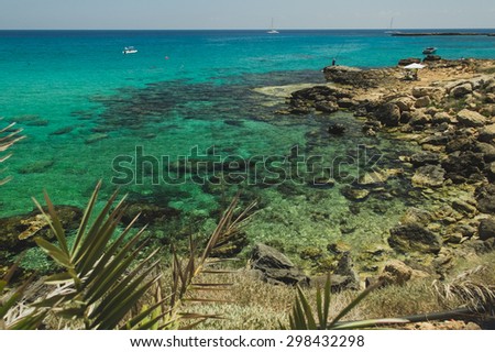 Rocky beach with tropical vegetation and crystal clear blue water with incidental people and yacht on a background, on the sunny day, Protaras in Cyprus