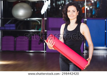 Waist up portrait of nice woman with curly short hair in sports wear holding pink yoga mat on a sports equipment background at the gym