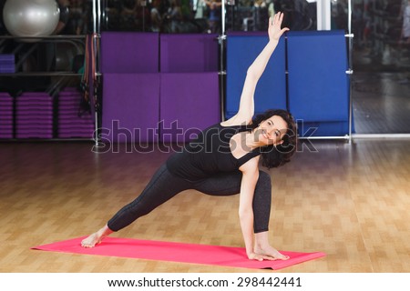 Happy athletic woman with curly hair wearing on black shirt and leggings posing in yoga asana on pink rubber mat at the gym