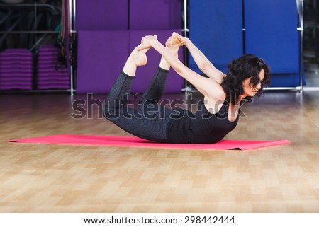 Active woman with curly hair wearing on black shirt and leggings make yoga stretching exercise on pink yoga mat on a sports equipment background at the gym