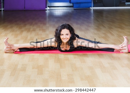 Athletic brunet woman with curly short hair wearing on black shirt and leggings make yoga stretching exercise on pink yoga mat on a sports equipment background at the gym