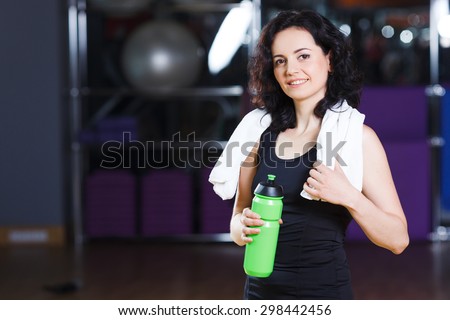 Waist up portrait of pretty woman with curly short hair in sports wear holding green bottle with towel on a sports equipment background at the gym