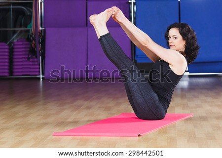 Fitness girl with curly hair wearing on black shirt and leggings put her legs up on pink yoga mat on a sports equipment background at the gym