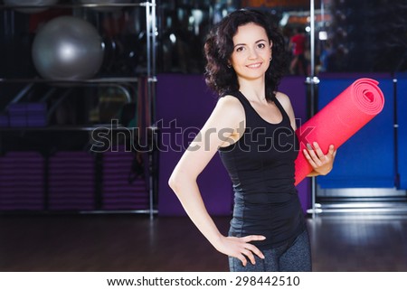 Waist up portrait of smiling fitness woman in sports wear holding pink yoga mat on a sports equipment background at the gym
