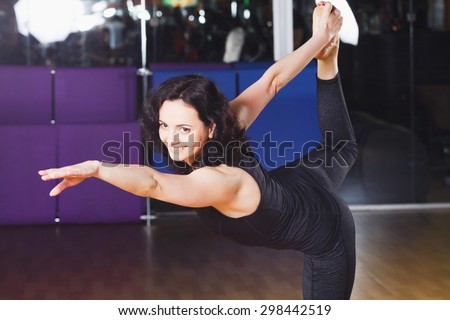 Brunet woman with curly hair wearing on black shirt and leggings stand in yoga asana - Dancer Pose on a sports equipment background at the gym