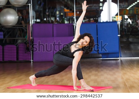 Sporty brunet woman with curly short hair wearing on black shirt and leggings make yoga stretching exercise on pink yoga mat on a sports equipment background at the gym