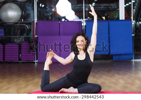 Active fitness woman with curly short hair wearing on black shirt and leggings make yoga stretching exercise and smiling on pink yoga mat on a sports equipment background at the gym