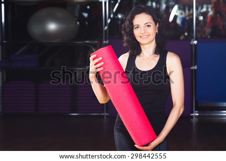 Waist up portrait of happy woman with curly short hair in sports wear holding pink yoga mat on a sports equipment background at the gym