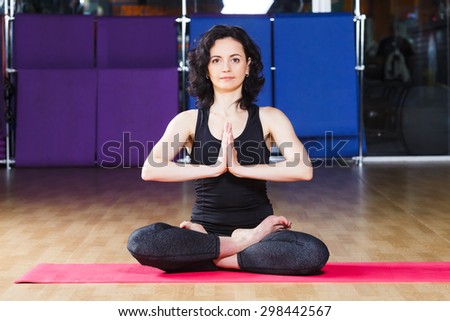 Active fitness woman with curly short hair wearing on black shirt and leggings is sitting in a lotus yoga pose on pink yoga mat on a sports equipment background at the gym