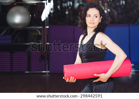 Waist up portrait of sporty woman with curly short hair in sports wear holding pink yoga mat on a sports equipment background at the gym