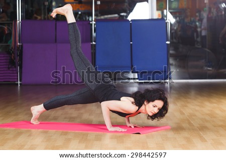 Sporty woman with dark curly hair wearing on black shirt and leggings make yoga stretching exercise on pink yoga mat on a sports equipment background at the gym