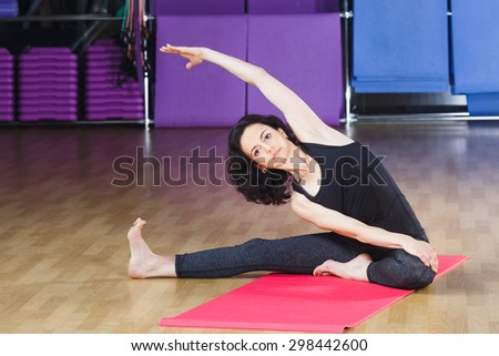 Pretty young woman with dark curly hair wearing on black shirt and leggings doing exercises on pink yoga mat on a sports equipment background at the gym