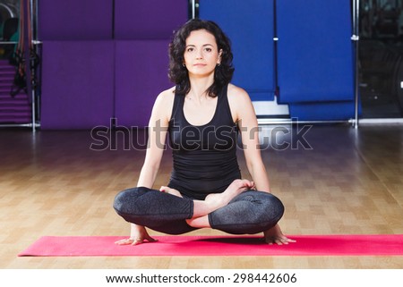 Beauty woman with curly short hair wearing on black shirt and leggings is meditating tolasana pose on pink yoga mat on a sports equipment background at the gym