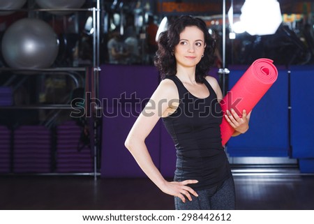 Waist up portrait of fitness woman in sports wear holding pink yoga mat on a sports equipment background at the gym