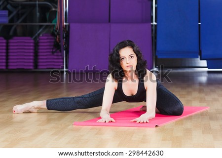 Brunet girl with curly hair wearing on black shirt and leggings doing exercises on pink yoga mat on a sports equipment background at the gym